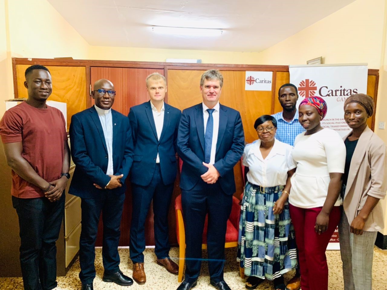 Group photo of CARITAS Gambia staff and international partners standing together, including a priest and diverse team members, in a professional setting with a CARITAS banner in the background.