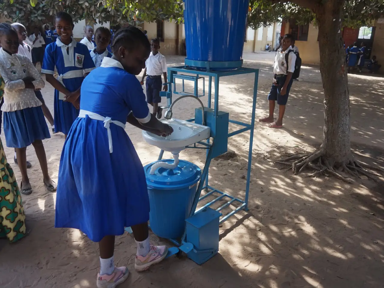 A young schoolgirl in a blue uniform washing her hands at a handwashing station, surrounded by other students, as part of a hygiene and sanitation initiative at a school in The Gambia.